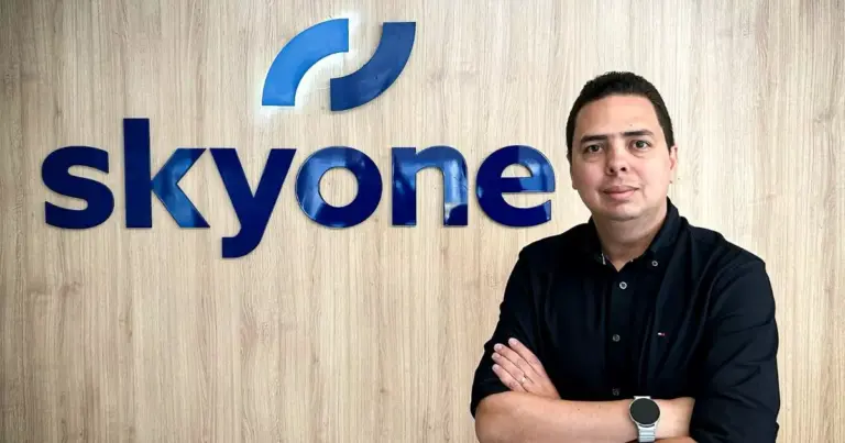 Profissional posando com braços cruzados em frente ao logotipo da Skyone em escritório moderno, destacando identidade corporativa e ambiente empresarial.