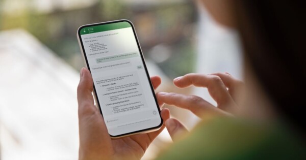 Uma pessoa segura um smartphone com uma mão enquanto usa o dedo da outra mão para interagir com a tela. O dispositivo está em posição vertical e exibe uma conversa em um aplicativo de mensagens com um assistente virtual chamado “Liza”. O cabeçalho da conversa aparece em verde, com o nome “Liza” no topo. Na tela, há uma mensagem inicial do assistente se apresentando e oferecendo ajuda com tarefas como informações e serviços. Mais abaixo, aparecem respostas em formato de lista, com opções numeradas e descrições de locais ou serviços, incluindo nomes e endereços. Na parte inferior, há um campo de texto indicando onde digitar uma resposta. O cenário ao redor está desfocado, sugerindo um ambiente interno próximo a uma janela ou varanda, com luz natural iluminando a cena. A pessoa e o celular estão em foco no primeiro plano, enquanto o fundo apresenta tons suaves e esverdeados, dando a impressão de vegetação ou paisagem externa ao longe (Localiza&Co)