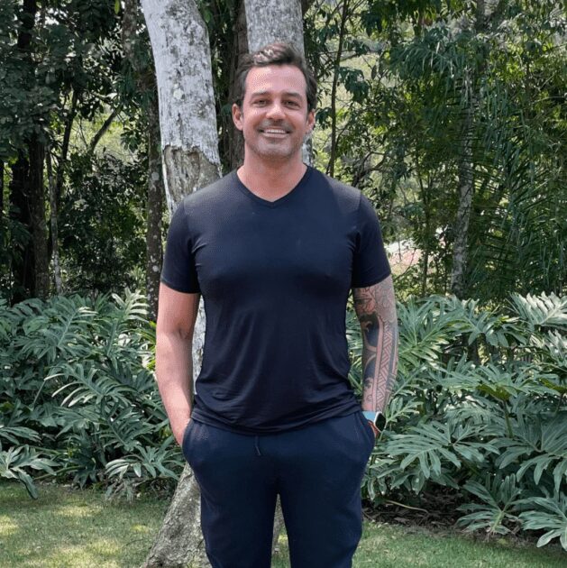 Foto de um homem sorridente vestindo uma camiseta preta e calça escura, com uma tatuagem no braço esquerdo. Ele está ao ar livre, em pé, com as mãos nos bolsos, em um ambiente de vegetação densa com árvores e plantas tropicais ao fundo.