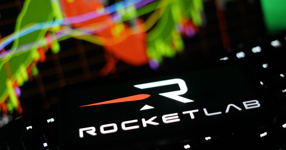 Logotipo da Rocket Lab exibido na tela de um smartphone, com gráficos financeiros coloridos desfocados ao fundo, representando investimentos, mercado de ações, tecnologia espacial e desempenho da empresa aeroespacial.