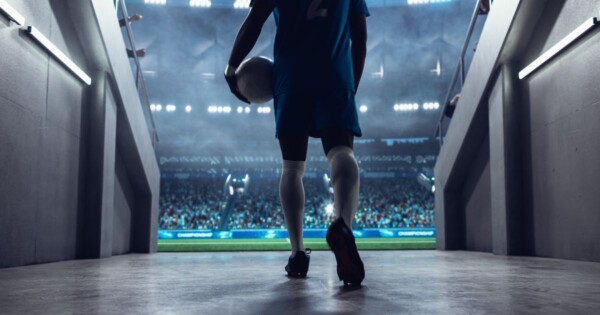 **Descrição alternativa (alt text):** Vista a partir de um túnel de acesso ao campo, mostrando uma pessoa vestindo uniforme esportivo azul caminhando em direção a um estádio iluminado, segurando uma bola de futebol sob o braço. A cena é capturada por trás, com foco nas pernas e nas chuteiras. O túnel possui paredes laterais com corrimãos e iluminação artificial. Ao fundo, um grande estádio aparece cheio de público, com luzes fortes e o campo verde visível na saída do túnel, criando um contraste entre a área interna sombreada e o ambiente externo luminoso. (Sport)