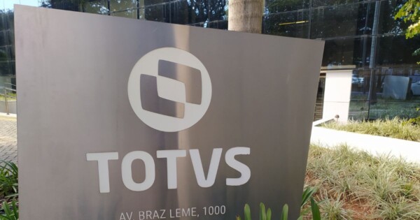 Placa metálica moderna com o logotipo da empresa TOTVS em destaque, composta por um símbolo circular e o nome “TOTVS” em letras grandes e brancas. A placa está instalada em área externa, cercada por jardim com plantas ornamentais e pedras. Ao fundo, há um prédio com fachada de vidro refletivo e um tronco de árvore ao lado da placa. Na parte inferior da placa, aparece parcialmente o endereço “AV. BRAZ LEME, 1000”. A iluminação é natural, sugerindo ambiente diurno. (Caruaru)