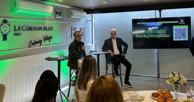 Ambiente interno de um evento ou palestra em um espaço elegante, com iluminação suave em tons de verde e branco. À esquerda, há uma parede com o logotipo “Le Cordon Bleu São Paulo – Culinary Village” iluminado em verde. Abaixo do logotipo, dois participantes estão sentados em banquetas altas, cada um ao lado de uma pequena mesa redonda com copos de água e garrafas. Um dos participantes segura um microfone e parece estar falando, enquanto o outro observa. Ambos usam roupas formais ou casuais elegantes. À frente deles, há uma pequena plateia sentada, vista de costas, acompanhando a conversa. À direita da cena, um monitor grande exibe uma apresentação com o texto: “Um evento digital. Resoluções infinitas.” Também aparecem os logotipos “zendesk relat[e]” e “Zendesk”, além de um QR code com a indicação “Acesse e compartilhe”. No primeiro plano, sobre uma mesa, há itens de coffee break, incluindo croissants, copos, pratos e um pequeno arranjo de flores brancas. Ao fundo, janelas com persianas fechadas e aparelhos de ar-condicionado instalados na parede completam o ambiente. (Motiva)