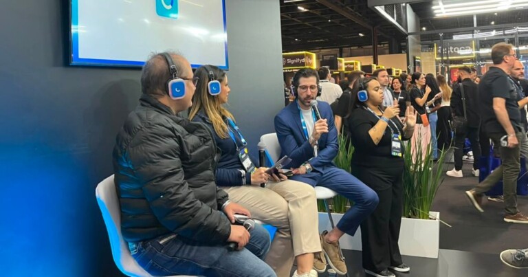 Painel de discussão realizado em um evento corporativo da Cielo. Três participantes estão sentados em bancos altos usando fones de ouvidodiante de um estande com iluminação azul. Uma das pessoas segura um microfone enquanto falae outra segura materiais de apoio. Ao lado do grupohá uma intérprete de Libras realizando a tradução da apresentação. Ao fundodiversos visitantes circulam pelos corredores do evento entre estandes iluminados e áreas de exposição. Vasos com plantas ornamentais decoram o espaçocontribuindo para a composição do ambiente. A imagem retrata uma apresentação ou conversa mediada em um ambiente de networking e tecnologia.