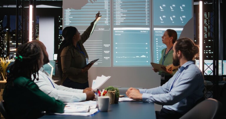 Equipe corporativa reunida em uma sala de reuniões analisando dashboards e indicadores de desempenho projetados em uma tela digital. A apresentação exibe gráficos, métricas financeiras e dados de negócios em um ambiente tecnológico moderno, voltado para análise estratégica, business intelligence e tomada de decisões. (salarial)