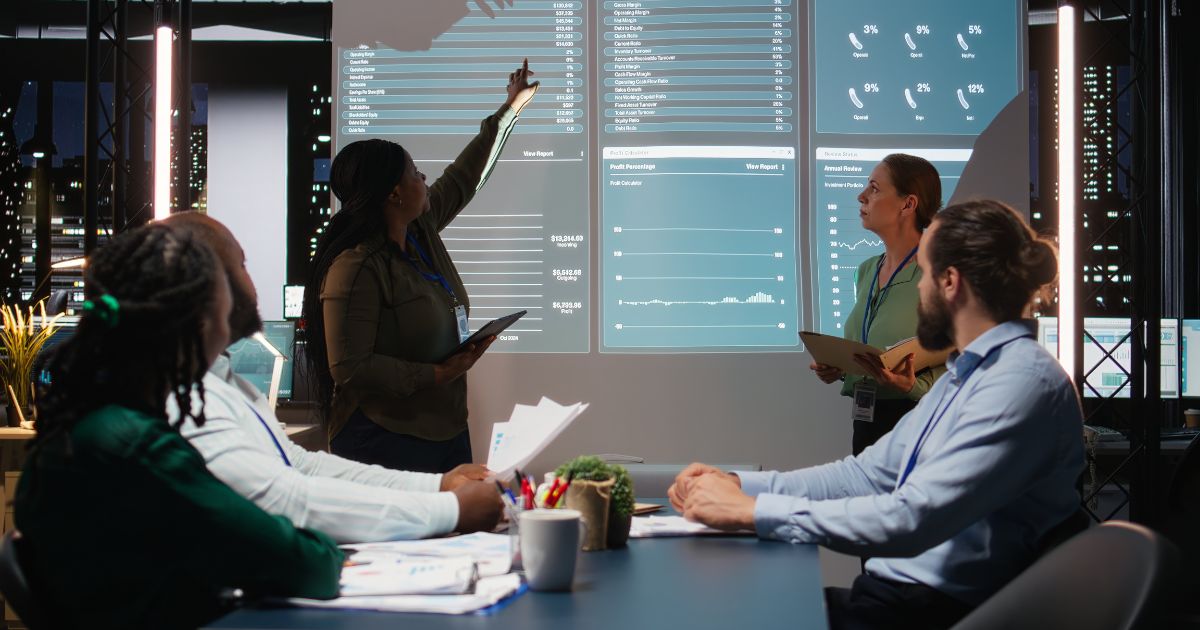 Equipe corporativa reunida em uma sala de reuniões analisando dashboards e indicadores de desempenho projetados em uma tela digital. A apresentação exibe gráficos, métricas financeiras e dados de negócios em um ambiente tecnológico moderno, voltado para análise estratégica, business intelligence e tomada de decisões. (salarial)