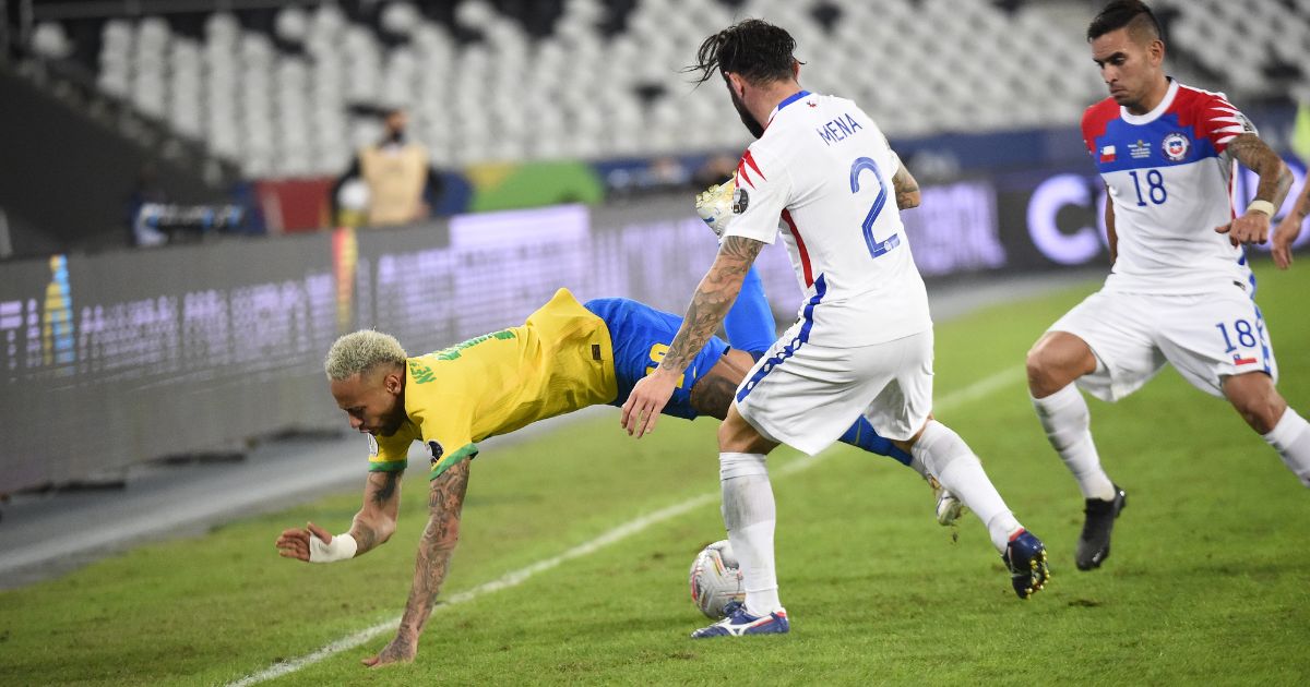 Lance de partida de futebol entre Brasil e Chile, com jogador da seleção brasileira caindo próximo à linha lateral enquanto atletas da equipe adversária disputam a bola em campo.