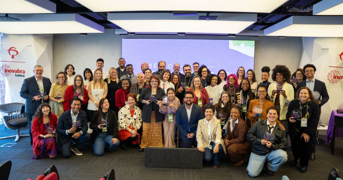 Descrição alternativa (alt text): Fotografia de grupo realizada em um auditório corporativo. Cerca de 40 pessoas estão reunidas em frente a um telão, organizadas em várias fileiras, algumas em pé e outras agachadas na parte frontal. Diversas pessoas seguram troféus transparentes de premiação. O ambiente possui iluminação moderna no teto, paredes claras e banners laterais com a marca “inovabra habitat”. Ao fundo, há uma tela em tons de lilás com elementos gráficos. As pessoas usam roupas variadas, incluindo trajes sociais, casuais e coloridos. A imagem registra um momento de reconhecimento ou encerramento de um evento, com todos posicionados para uma foto oficial de grupo. (futuro)