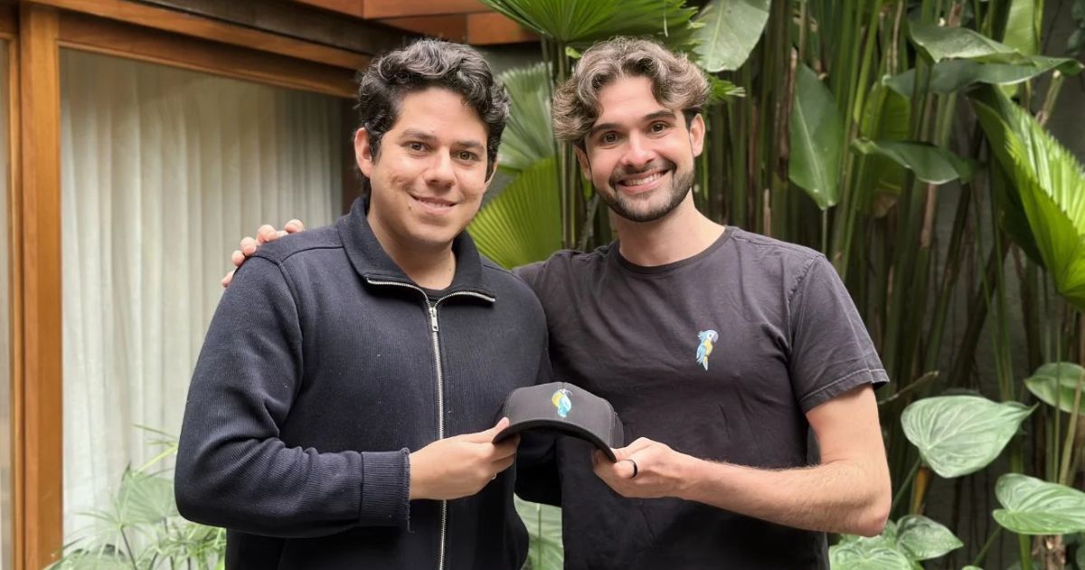 Duas pessoas posam lado a lado em uma área externa cercada por vegetação tropical. Ambas estão voltadas para a câmera e seguram juntas um boné escuro com um pequeno logotipo colorido na parte frontal. Uma das pessoas veste uma jaqueta escura com zíper, enquanto a outra usa camiseta escura com o mesmo logotipo estampado no peito. Ao fundo, há plantas de grande porte e parte de uma estrutura com esquadrias de madeira e cortinas claras. (Nero.AI)