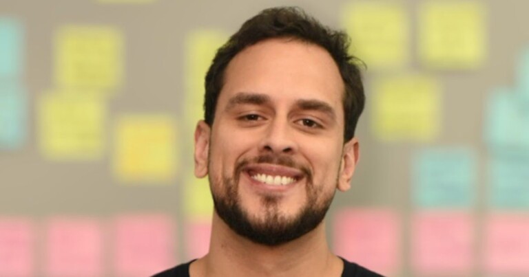 Retrato em close de uma pessoa posicionada ao centro da imagem, olhando para a câmera. A pessoa possui barba curta e usa uma camiseta escura. Ao fundo, desfocado, há uma parede com diversos blocos de notas adesivas em tons de amarelo, azul e rosa, organizados em diferentes áreas. A iluminação é suave e o foco está no rosto da pessoa retratada. (inteligência)