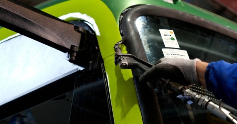 Detalhe da manutenção e instalação de vidro automotivo em ônibus Marcopolo, com técnico utilizando ferramenta especializada para ajustar o para-brisa em ambiente industrial.