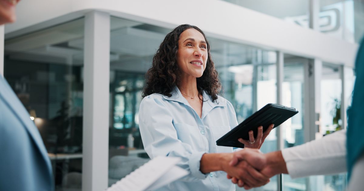 Pessoa em ambiente corporativo moderno cumprimenta outra pessoa com um aperto de mão enquanto segura um tablet. Ao fundo, há divisórias de vidro e escritórios iluminados, compondo um cenário profissional de reunião ou networking. (sonda)