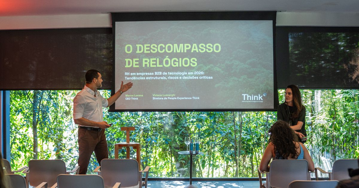 Sala de eventos com cadeiras voltadas para um palco. Uma pessoa em pé apresenta um slide projetado com o título “O Descompasso de Relógios: RH em empresas B2B de tecnologia em 2026: Tendências estruturais, riscos e decisões críticas”. Outra pessoa está ao lado da apresentação. Ao fundo, grandes janelas revelam vegetação densa, trazendo bastante luz natural ao ambiente. Alguns participantes aparecem sentados assistindo à palestra. (capital)