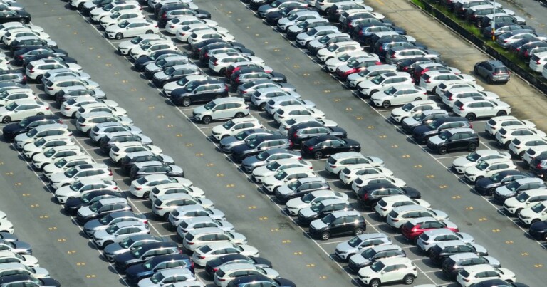 Vista aérea de um grande pátio de estacionamento com centenas de automóveis organizados em longas fileiras paralelas. Os veículospredominantemente nas cores brancapretacinza e prataocupam quase todas as vagas demarcadas no piso asfaltado. Entre as fileiras há corredores largos para circulaçãocom numeração pintada no chão indicando as posições de estacionamento. A imagem destaca a alta concentração de veículos e a organização do espaçoremetendo a um centro de distribuiçãoporto automotivo ou grande estacionamento do setor automotivo. (crescimento)
