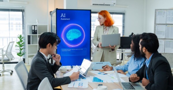 Equipe reunida em sala de reuniões corporativa analisando soluções de inteligência artificial, com apresentação de assistente virtual em tela digital e notebooks sobre a mesa.