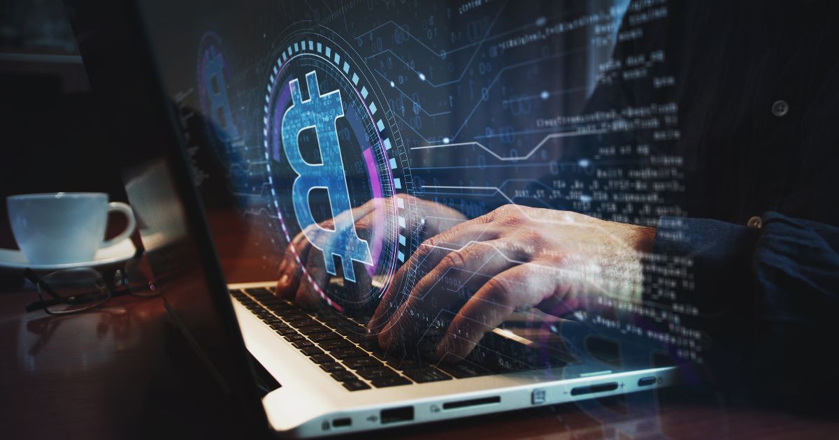 Evertec conclui aquisição da BBChain 2 Imagem conceitual relacionada a criptomoedas, tecnologia financeira e segurança digital. Em primeiro plano, mãos digitam em um computador portátil sobre uma mesa escura. À esquerda da cena, há uma xícara branca e um par de óculos apoiados sobre a superfície. Sobreposta à imagem aparece uma interface digital com elementos gráficos em tons de azul e roxo, incluindo o símbolo do Bitcoin destacado no centro. Linhas de conexão, códigos, números binários e blocos de dados espalham-se pela composição, simulando uma rede digital. A iluminação é baixa, com reflexos de luz azulada realçando os elementos tecnológicos. A imagem representa temas como blockchain, transações digitais, criptomoedas e cibersegurança. (Evertec)