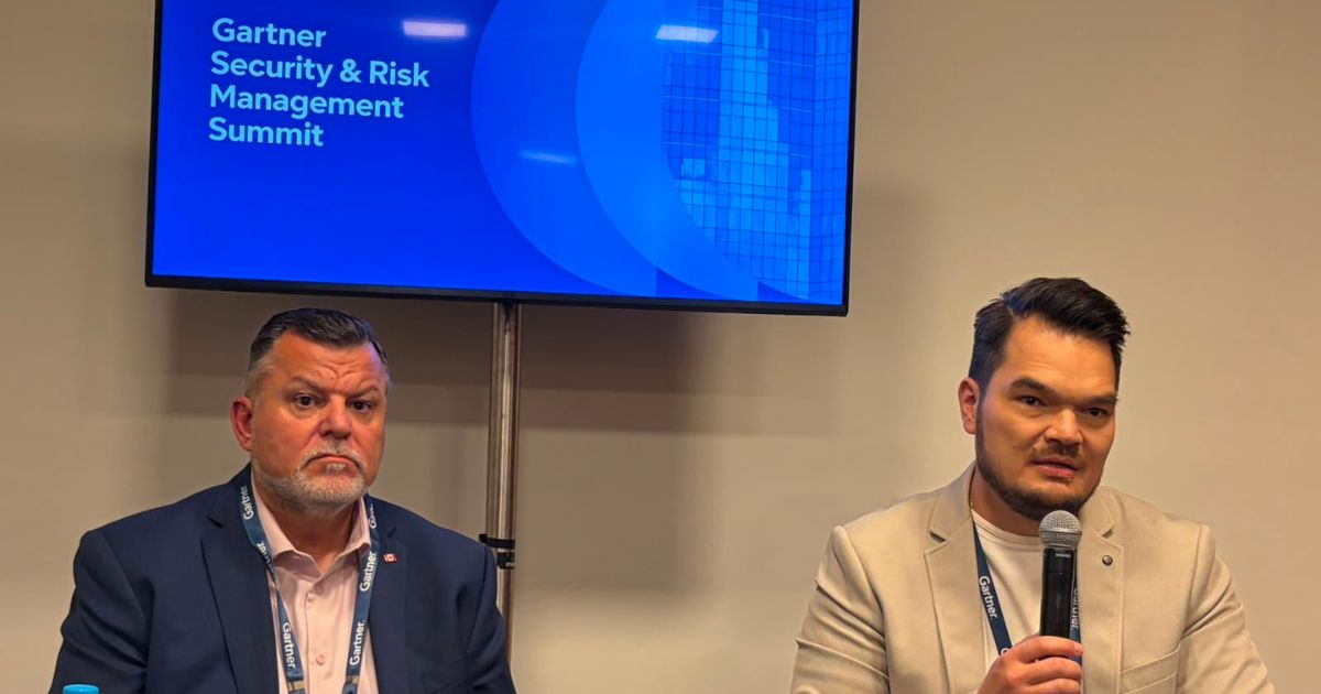 Investimentos crescentes em IA exigem nova postura dos CISOs, afirma Gartner 2 Fotografia de um painel de discussão em evento sobre segurança da informação e gestão de riscos. Duas pessoas estão sentadas lado a lado em um ambiente interno, utilizando crachás de identificação. A pessoa à direita segura um microfone durante a apresentação. Ao fundo, um monitor exibe o texto “Gartner Security & Risk Management Summit” sobre um fundo azul com elementos gráficos circulares. O espaço possui paredes claras e iluminação uniforme, característica de salas de conferência e eventos corporativos. A composição destaca a participação dos palestrantes em uma sessão de debate ou compartilhamento de conhecimento relacionada à cibersegurança e gestão de riscos.