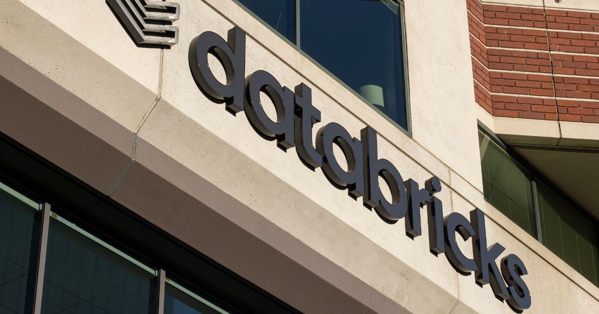 Databricks conclui aquisição da Panther 2 Fotografia da fachada de um edifício comercial com o logotipo e o nome “databricks” em letras pretas fixadas na parede externa. A construção combina revestimento claro, janelas amplas de vidro e uma seção em tijolos aparentes na parte superior direita. A imagem foi capturada em ângulo inclinado, destacando a sinalização da empresa ao longo da fachada. A iluminação natural evidencia as texturas dos materiais e os contornos das letras. O cenário remete a um ambiente corporativo e tecnológico, com foco na identidade visual da Databricks e na arquitetura do edifício.