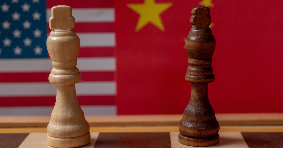 EUA preparam restrição a componentes chineses para data centers em nova frente da disputa tecnológica 4 Imagem conceitual que representa a rivalidade estratégica entre Estados Unidos e China. Em primeiro plano, sobre um tabuleiro de xadrez, estão posicionadas duas peças do tipo rei: uma peça clara à esquerda e uma peça escura à direita. Ao fundo, aparecem desfocadas as bandeiras dos Estados Unidos e da China, preenchendo grande parte da composição. As cores predominantes são vermelho, azul, branco e amarelo, associadas às bandeiras dos dois países. O enquadramento destaca as peças de xadrez como símbolo de competição, negociações e disputas geopolíticas entre as duas potências. (chineses)