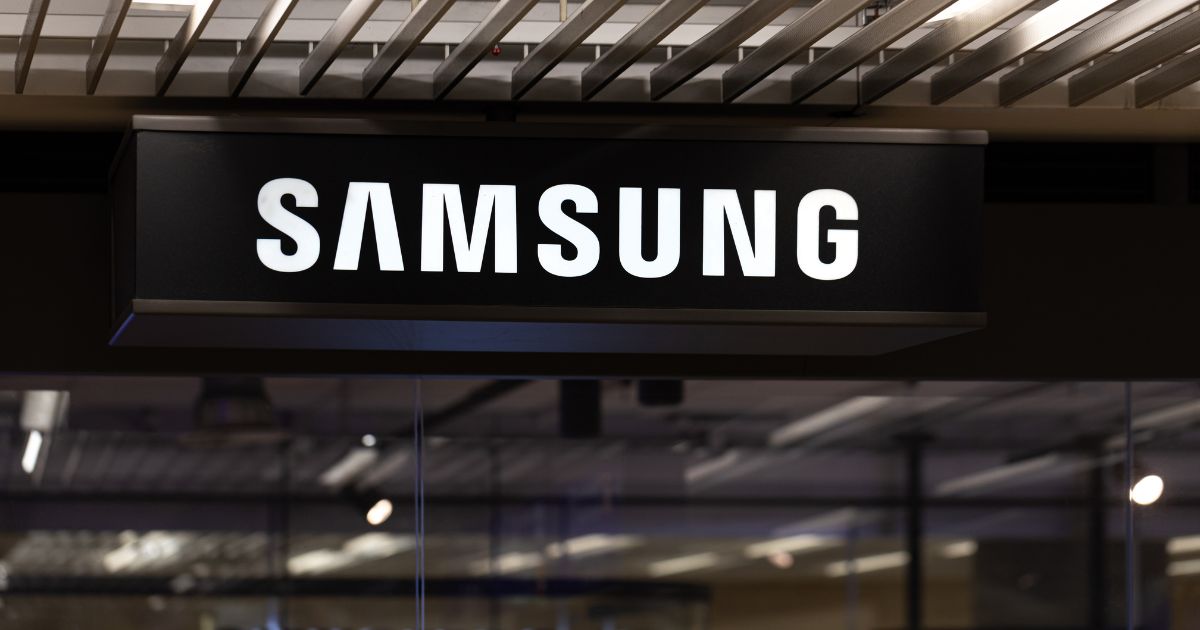 Investidores de varejo pressionam Samsung por recompra de ações e assembleia extraordinária 6 Fotografia de um letreiro iluminado da Samsung instalado em ambiente interno. O nome da marca aparece em letras brancas de grande porte sobre uma estrutura retangular preta suspensa. Acima do letreiro, há um teto com vigas metálicas paralelas e iluminação embutida. Na parte inferior da imagem, são visíveis superfícies de vidro e elementos arquitetônicos desfocados, sugerindo um espaço comercial, showroom ou estande de exposição. A composição destaca a identidade visual da Samsung em um ambiente moderno e corporativo.