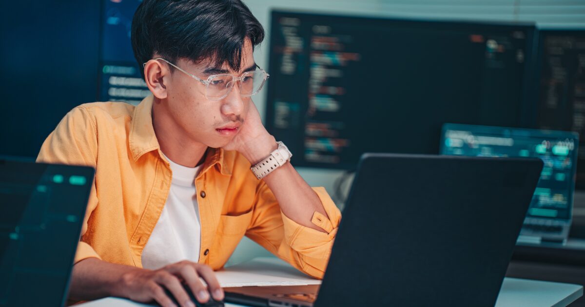 IA eleva régua para juniores em vagas de tecnologia, mostra Gupy 2 Profissional de tecnologia trabalha em notebook diante de telas com códigos de programação, em cenário que representa as novas exigências para desenvolvedores em meio ao avanço da inteligência artificial. (juniores)