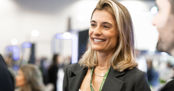 Lara Paiva, nova vice-presidente de Estratégia em Saúde e Expansão da Telepatia, aparece sorrindo durante um evento, vestindo blazer cinza-escuro, blusa branca, colares e um cordão de identificação verde da empresa. Ao fundo, aparecem participantes e estruturas do evento desfocados. A executiva assume a estratégia de expansão da healthtech e a coordenação das iniciativas de governança clínica e inteligência artificial aplicada à saúde. (Lara Paiva, Telepatia, Liderança, Healthtech, Inteligência Artificial, Saúde Digital)