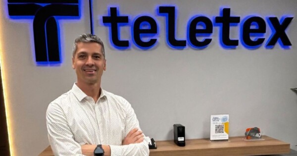 Sérgio Milani, novo Head de Utilities da Teletex, posa para foto institucional com camisa branca estampada e braços cruzados, diante do logotipo da companhia iluminado em azul. O executivo assume a liderança da vertical de energia e infraestrutura crítica, com foco em tecnologia operacional, telecomunicações e cibersegurança. (Sérgio Milani, Teletex, Utilities, Tecnologia, Cibersegurança)